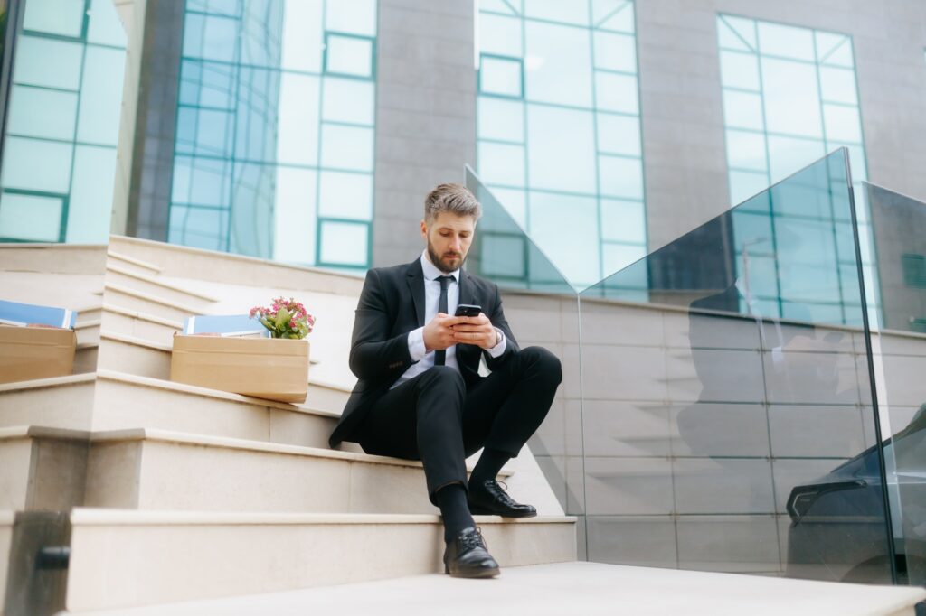 Sad laid-off man in suit checks phone after job loss on office stairs